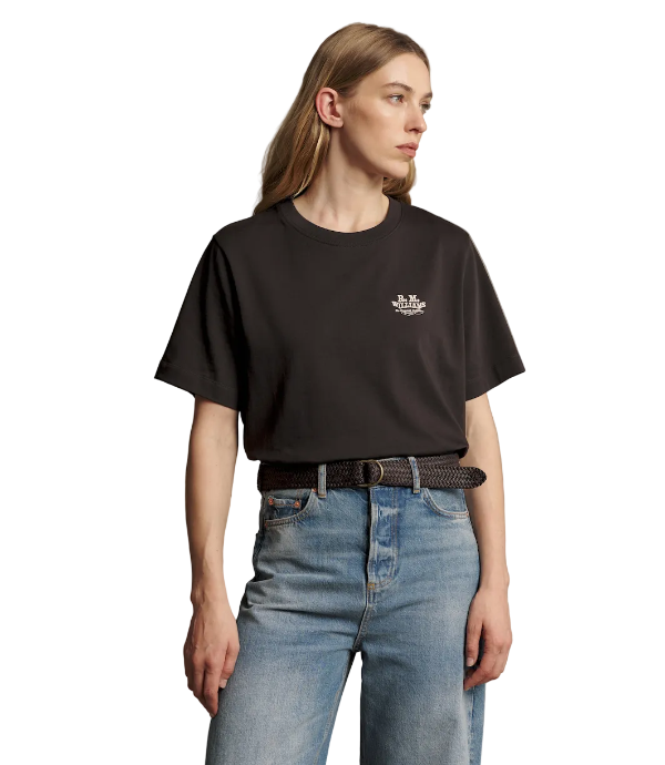 A woman wearing a black short-sleeve crewneck t-shirt with a small white embroidered logo on the left chest, paired with blue jeans and a black belt, against a solid taupe background.