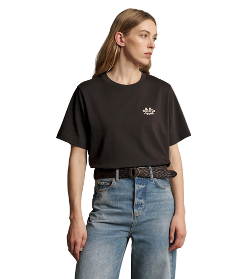 A woman wearing a black short-sleeve crewneck t-shirt with a small white embroidered logo on the left chest, paired with blue jeans and a black belt, against a solid taupe background.