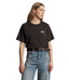 A woman wearing a black short-sleeve crewneck t-shirt with a small white embroidered logo on the left chest, paired with blue jeans and a black belt, against a solid taupe background.