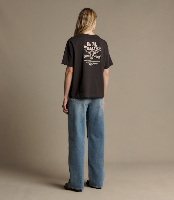 A woman stands with her back to the camera wearing a black t-shirt with a large white graphic print on the back and wide-leg blue jeans, photographed in a studio setting with a solid beige background.