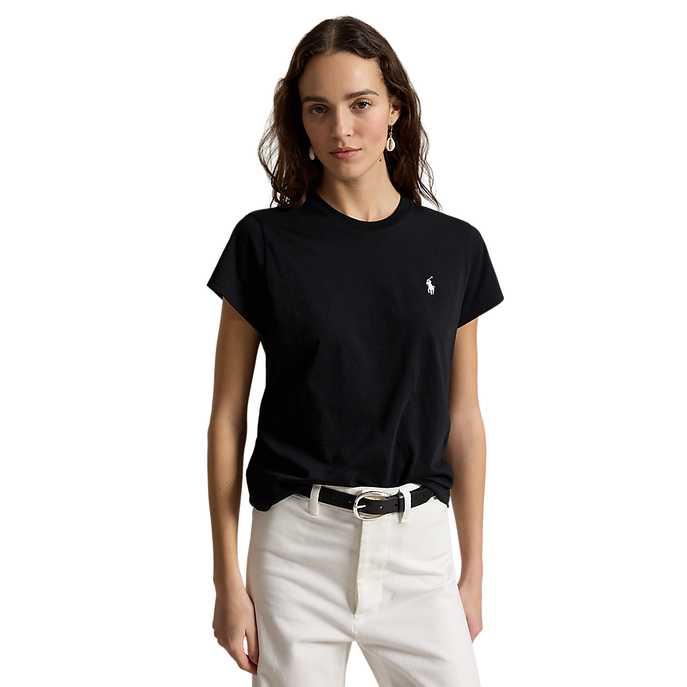 A woman modeling a black short-sleeve crewneck t-shirt with a small white embroidered polo player logo on the left chest, paired with white pants and a black belt, against a plain white background.