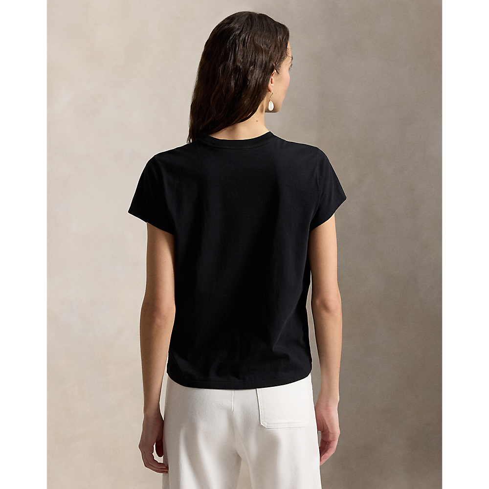 A woman shown from the back wearing a plain black short-sleeve crewneck T-shirt and off-white trousers, standing against a textured beige studio backdrop.