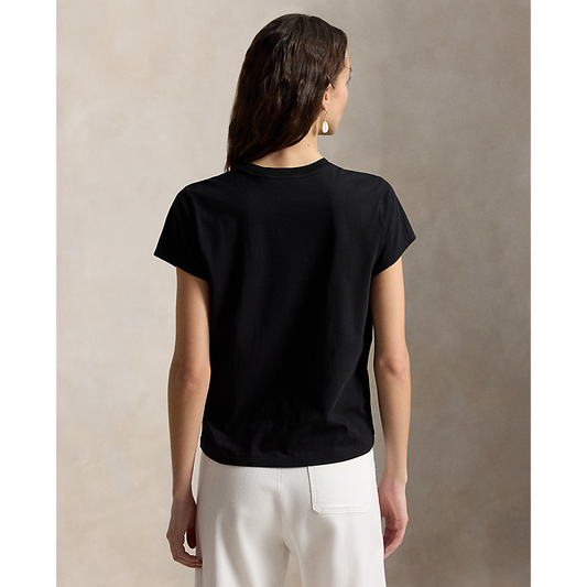 A woman shown from the back wearing a plain black short-sleeve crewneck T-shirt and off-white trousers, standing against a textured beige studio backdrop.