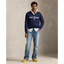 A male model stands against a textured beige studio backdrop wearing a navy blue Polo Sport quarter-zip sweatshirt over a white collared shirt, blue jeans, and yellow shoes. The Polo Sport logo is clearly visible on the chest.