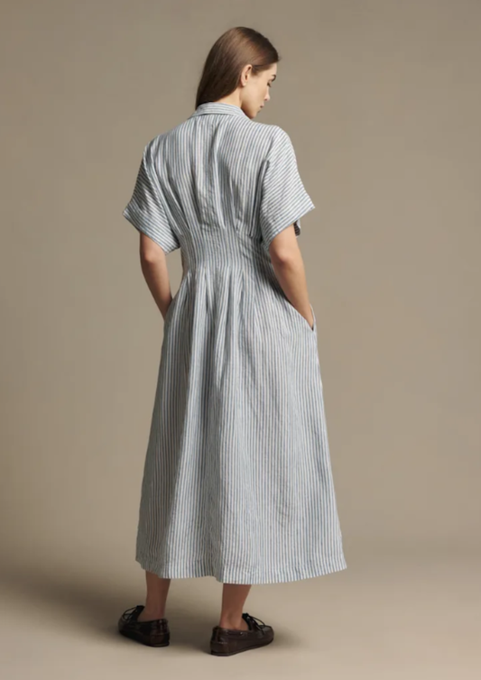 A woman viewed from the back wearing a light blue and white vertically striped midi shirt dress with short sleeves and side pockets, paired with dark brown loafers, against a solid beige studio background.