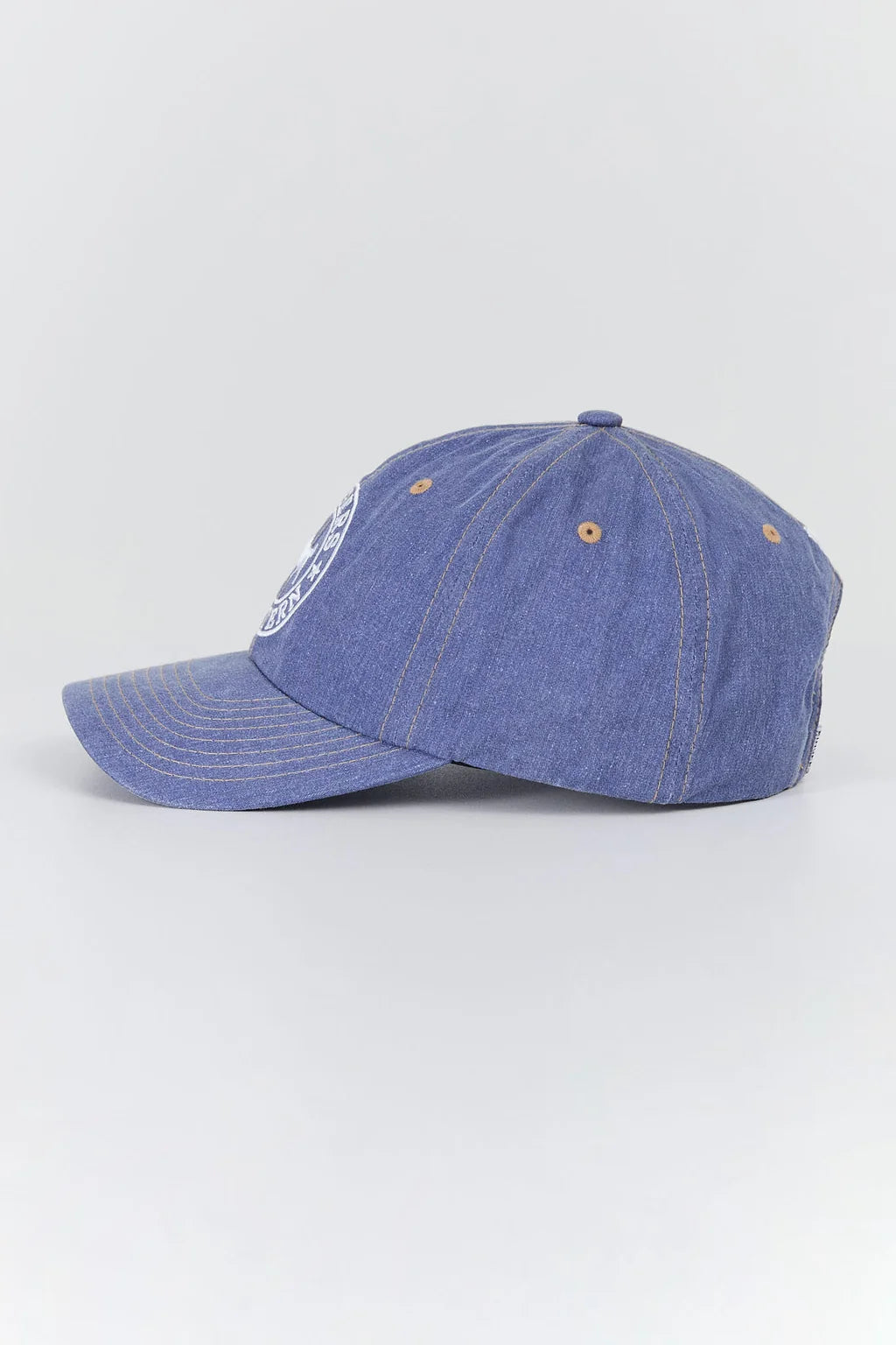 A blue denim baseball cap with a curved brim, featuring a white embroidered logo on the side and orange eyelets, photographed on a light gray background.
