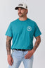 A man wearing a teal short-sleeve t-shirt with a circular white chest graphic, paired with light blue jeans and a white baseball cap, standing against a light gray studio background.