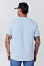 A man viewed from behind wearing a light blue crewneck t-shirt, blue jeans, and a black baseball cap, set against a plain light gray studio background.
