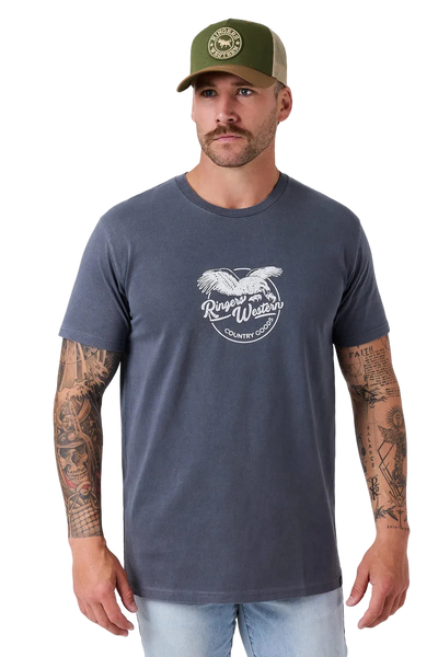 A man wearing a dark slate blue t-shirt with a white graphic print of an eagle and the words 'Ranger Wear Country Club'. He is also wearing an olive green trucker hat and light wash jeans, standing against a light gray studio background.