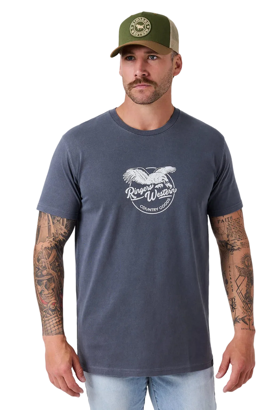 A man wearing a dark slate blue t-shirt with a white graphic print of an eagle and the words 'Ranger Wear Country Club'. He is also wearing an olive green trucker hat and light wash jeans, standing against a light gray studio background.