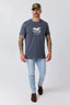 A man modeling a dark slate blue t-shirt with a white graphic on the chest, paired with light blue jeans, brown boots, and an olive green baseball cap, against a light gray studio background.