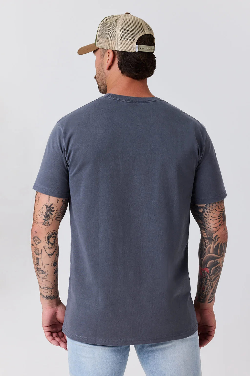 A man viewed from the back wearing a faded dark slate blue t-shirt, a beige trucker hat, and light blue jeans against a light gray studio background.