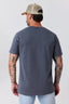 A man viewed from the back wearing a faded dark slate blue t-shirt, a beige trucker hat, and light blue jeans against a light gray studio background.