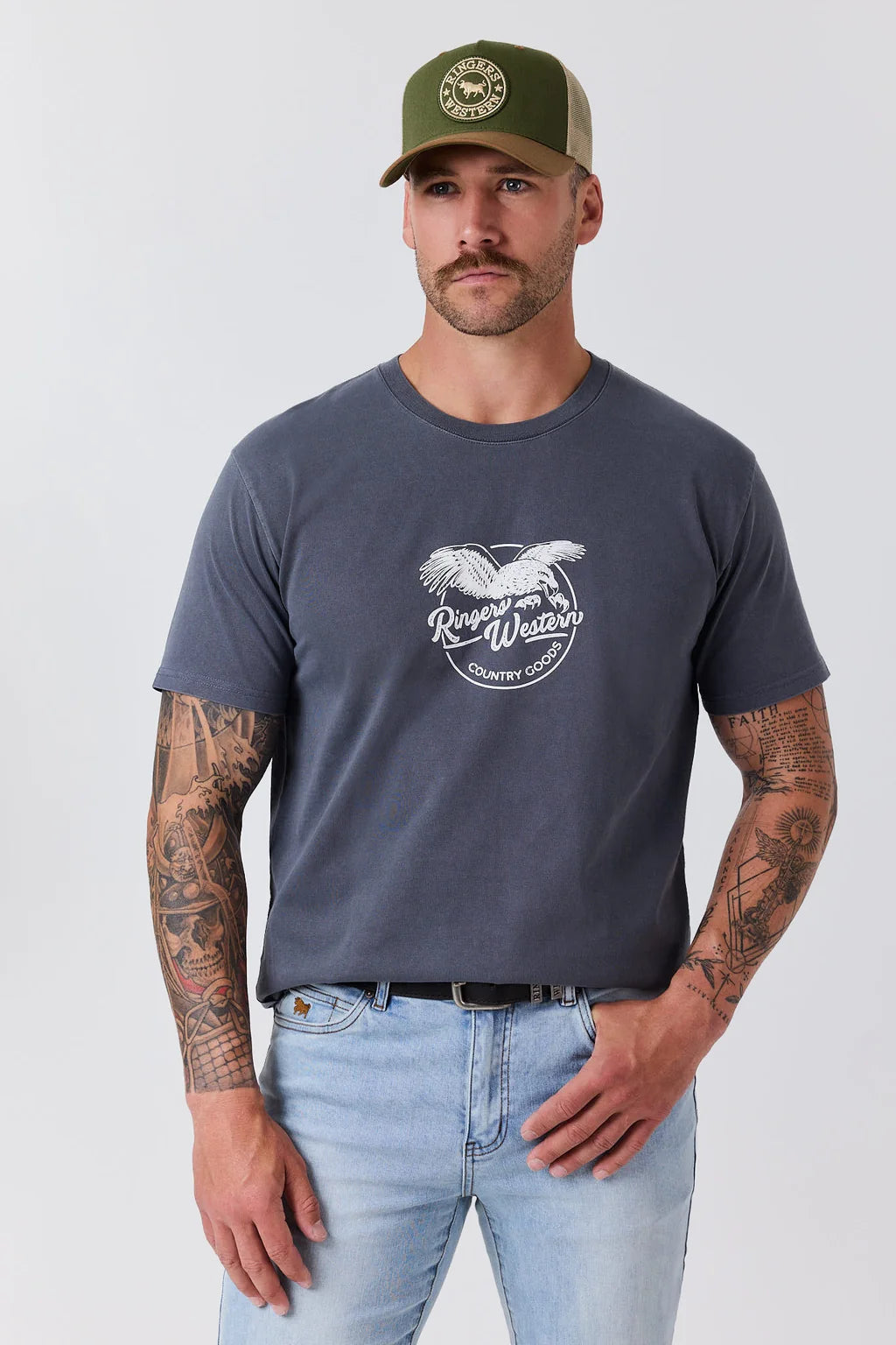 A man wearing a dark gray graphic t-shirt with a white horse logo and the words 'Rough Weather', paired with light blue jeans and an olive green trucker hat, standing against a light gray background.