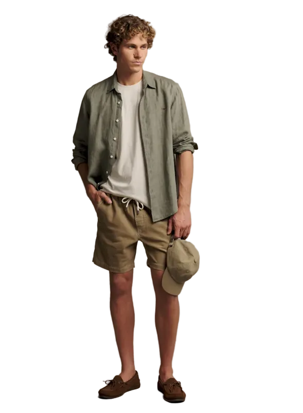 A man modeling a casual summer outfit consisting of a light sage green linen button-up shirt over a white t-shirt, khaki shorts, and brown suede loafers, holding a matching khaki cap against a solid beige studio background.