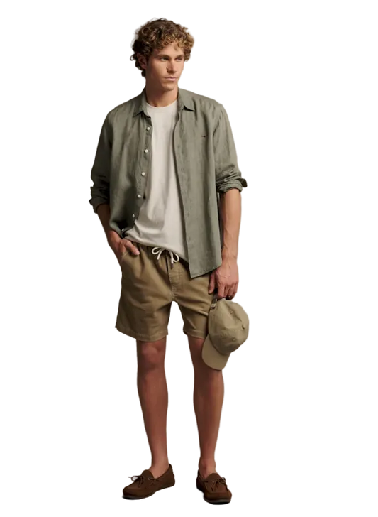 A man modeling a casual summer outfit consisting of a light sage green linen button-up shirt over a white t-shirt, khaki shorts, and brown suede loafers, holding a matching khaki cap against a solid beige studio background.