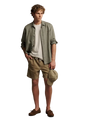A man modeling a casual summer outfit consisting of a light sage green linen button-up shirt over a white t-shirt, khaki shorts, and brown suede loafers, holding a matching khaki cap against a solid beige studio background.