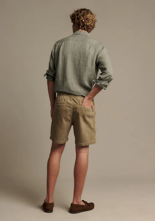 A man viewed from behind wearing a light sage green linen shirt, khaki linen shorts, and brown leather loafers, standing against a solid beige studio background.