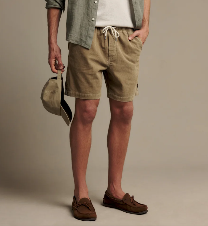 A man wearing khaki drawstring shorts, a light green shirt, a white t-shirt, and brown suede loafers, holding a beige baseball cap against a solid beige studio background.