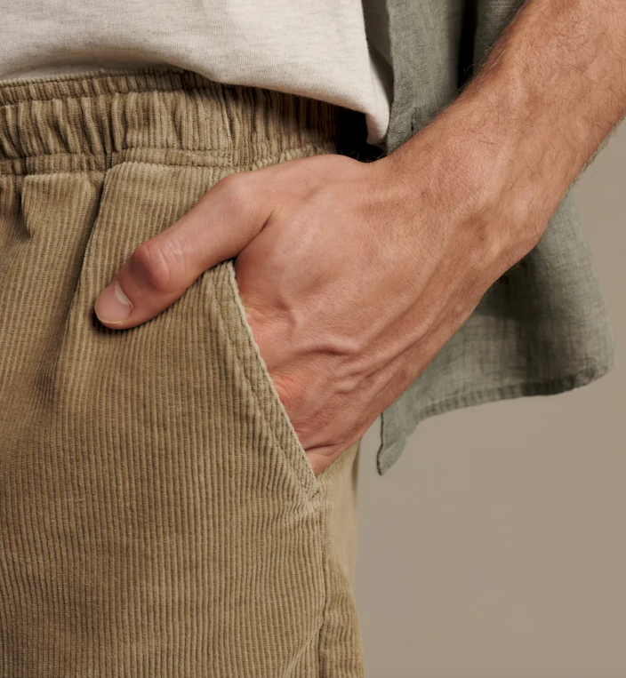 Close-up of a person wearing beige corduroy pants with a hand in the pocket, paired with a light gray shirt and a muted green draped fabric.