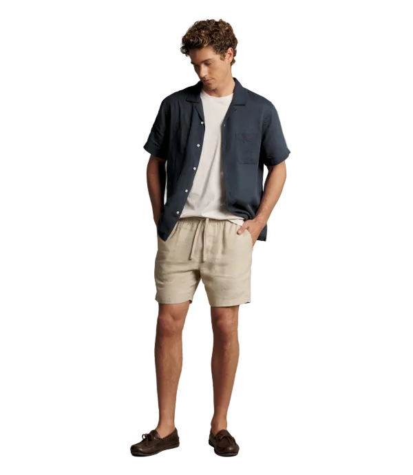 A man modeling a casual summer outfit consisting of a navy blue short-sleeve button-up shirt, a white crewneck t-shirt, beige linen shorts, and dark brown leather loafers, set against a solid warm gray studio background.