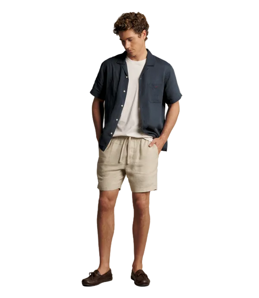 A man modeling a casual summer outfit consisting of a navy blue short-sleeve button-up shirt, a white crewneck t-shirt, beige linen shorts, and dark brown leather loafers, set against a solid warm gray studio background.