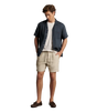 A man modeling a casual summer outfit consisting of a navy blue short-sleeve button-up shirt, a white crewneck t-shirt, beige linen shorts, and dark brown leather loafers, set against a solid warm gray studio background.