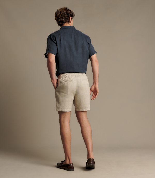 A man is shown from behind wearing a navy blue short-sleeve button-down shirt and light beige linen shorts, paired with dark brown leather loafers, against a solid warm gray studio background.