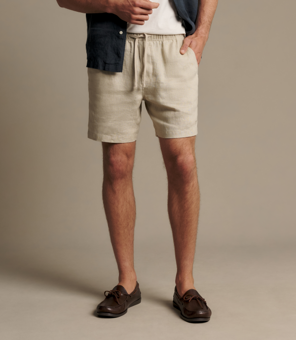 A man wearing beige linen drawstring shorts, styled with a white t-shirt, a dark blue overshirt, and brown leather loafers, standing against a solid beige studio background.