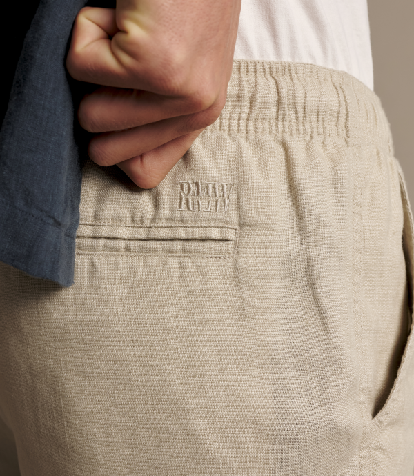 Close-up of a person wearing light beige textured trousers, showing the back pocket with an embroidered Tommy Hilfiger logo and a hand pulling up a dark blue shirt.