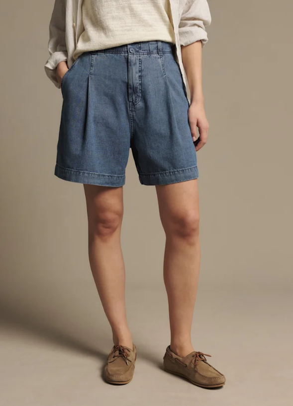 A model wearing blue denim shorts with pleats, a cream-colored top, and brown suede shoes, standing against a solid beige studio background.