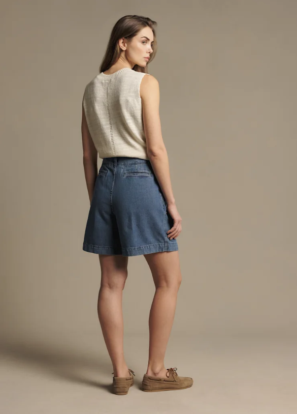 A woman is shown from the back wearing a sleeveless cream knit top, blue denim shorts, and brown boat shoes against a solid beige studio background.