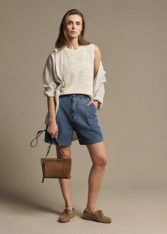A woman modeling a cream textured sleeveless top with long sleeves, blue denim shorts, brown suede shoes, and holding a brown leather handbag against a solid beige studio background.