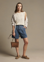 A woman modeling a cream textured sleeveless top with long sleeves, blue denim shorts, brown suede shoes, and holding a brown leather handbag against a solid beige studio background.