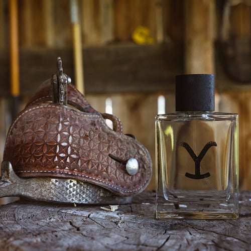 Ariat - Yellowstone - Eau de Cologne bottle beside a brown leather saddle, showcasing cowboy-inspired fragrance.
