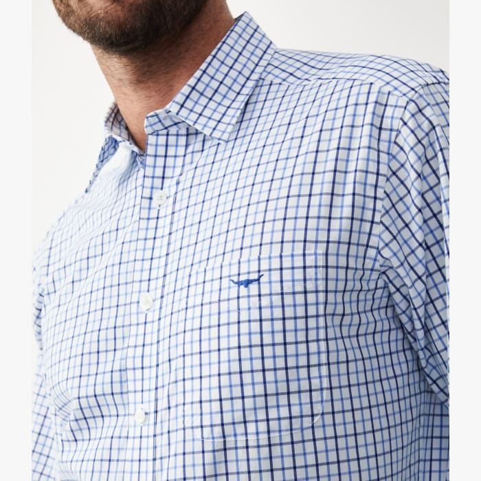 A close-up of a man wearing a pale blue and white checkered shirt with a point collar and long sleeves. The shirt features a chest pocket with embroidery and branded buttons.